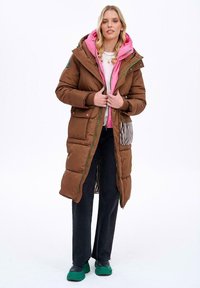 Długi brązowy puchowy płaszcz z różową kapturem, z zielonymi akcentami zamka błyskawicznego i paskowanymi bocznymi kieszeniami. Noszony z czarnymi spodniami i zielonymi butami.