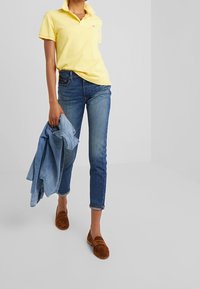 Žluté polo tričko, modré džíny a hnědé semišové loafers. Model drží světle modrou denimovou bundu. Jednoduchý design, volný střih a vyhrnuté džíny.