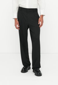 Pantalones negros de corte ajustado con una textura suave, que cuentan con un cierre de botón y una parte delantera plisada. Llevados con zapatos negros y una camisa blanca.
