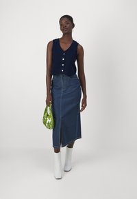 Ναυτική πλεκτή αμάνικη μπλούζα με κουμπιά, συνδυασμένη με τζιν φούστα με μπροστινή σχισμή. Λευκές μπότες μέχρι τον αστράγαλο και πράσινη τσάντα.