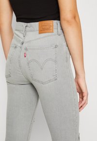 Persona que lleva jeans Levi's de color gris claro con bolsillos traseros visibles, etiqueta roja de Levi's y parche de cuero de la marca, combinado con una camiseta negra.