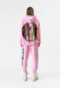 Sudadera rosa con alas de ángel de lentejuelas coloridas en la espalda, combinada con pantalones de chándal rosa a juego decorados con acentos de plumas blancas.