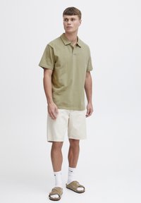 Olivengrønn polo-skjorte med krage, korte ermer, kombinert med hvite shorts og brune sandaler. Enkel, uformell design.