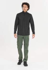 Schwarz langärmliges Zip-Up-Oberteil mit hohem Kragen, kombiniert mit grünen Cargo-Hosen mit Seitentaschen und schwarzen Wanderschuhen.