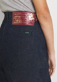 Mörkblå denimjeans har en röd läderlapp med en tecknad figur och text, samt en grön "LEVI'S" etikett på bakfickan.