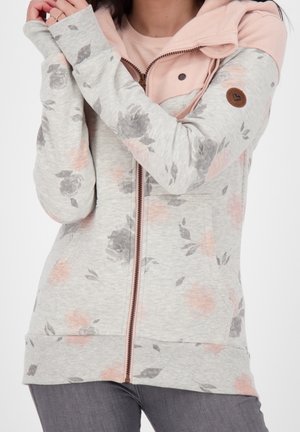 Femme portant un sweat à capuche zippé gris clair à motif floral avec une capuche et des poignets roses, croisant les bras près de son visage.