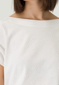 Hvid strikket top med en bred udskæring og korte ærmer, prydet med hævede blomster mønstre i stoffet for tekstur.