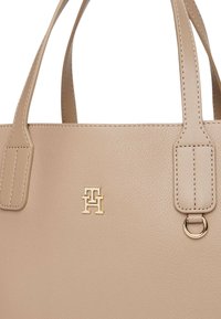 Beige leren draagtas met twee handvatten aan de bovenkant, voorzien van een gouden logo en stiksels. Gladde textuur met een gestructureerde vorm.