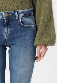 Blå jeans med hög midja, lätt urtvättade vid låren, kombinerade med en grön texturerad tröja med utsvängda ärmar.