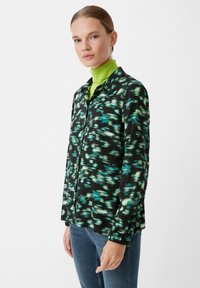 Black shirt with green and turquoise abstract pattern, collared, long sleeves, single chest pocket, paired with a bright green turtleneck.
