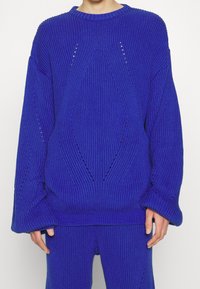 Pull bleu tricoté à coupe ample, avec motifs en losange et détails en maille ouverte. Poignets et encolure côtelés. Tissu doux et texturé.