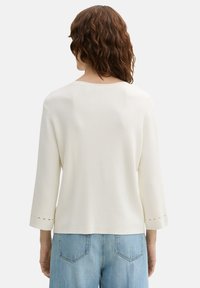 Witte trui met driekwartmouwen, gestructureerde stof, ronde halslijn en splitjes aan de mouwuiteinden, gecombineerd met lichtblauwe jeans.