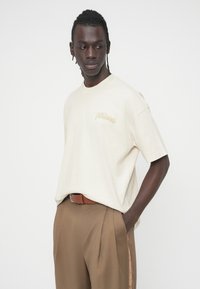 Μπεζ βαμβακερό T-shirt με κοντά μανίκια, με ένα λογότυπο σε καφέ απόχρωση στο στήθος, σε συνδυασμό με καφέ πλισέ παντελόνια και καφέ ζώνη.