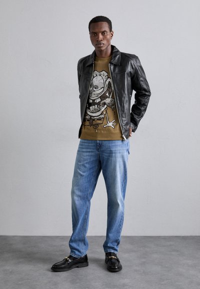 Sort læderjakke over en grafisk sweater i olivengrøn, blå jeans og sorte loafers med guld accenter. Afslappet outfit med klare lag.