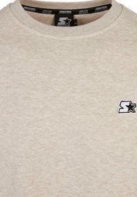 Sweat-shirt beige à col rond avec une petite étiquette logo Starter en noir et blanc sur la poitrine gauche et des étiquettes de marque à l'intérieur du col.