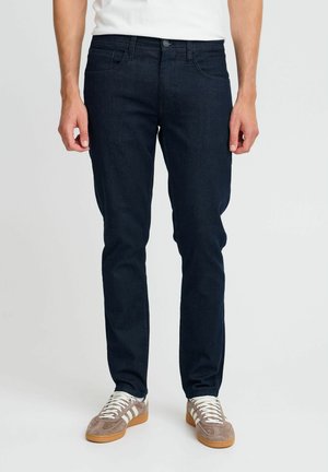 Dunkelblaue Denim-Jeans mit schlankem Schnitt, klassischem Fünf-Taschen-Design und Knopfverschluss. Getragen mit hellen Sneakers.