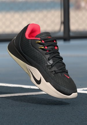 Chaussure de sport noire avec un dessus texturé, accents roses, œillets dorés et une semelle intermédiaire blanche arborant un logo noir, flottant au-dessus d'un terrain bleu.