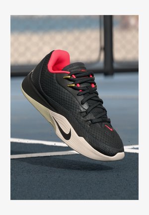Schwarzer Sportschuh mit strukturiertem Obermaterial, pinken Akzenten, goldenen Ösen und einer weißen Zwischensohle mit einem schwarzen Logo, schwebend über einem blauen Spielfeld.