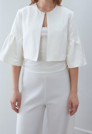 Femme portant une veste blanche courte avec de larges manches trois-quarts froncées, portée par-dessus un top blanc et un pantalon blanc taille haute.