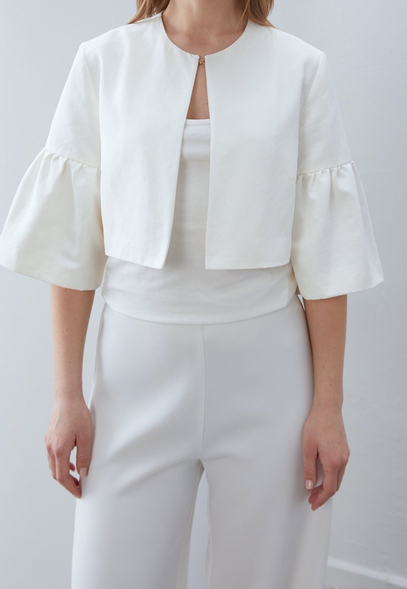 Femme portant une veste blanche courte avec de larges manches trois-quarts froncées, portée par-dessus un top blanc et un pantalon blanc taille haute.