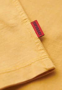 Zbliżenie na żółtą tkaninę z czerwoną metką z napisem „SUPERDRY” wszytą na krawędzi złożonego szwu ubrania.