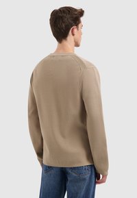 Man met zijn rug naar de camera, gekleed in een beige trui met lange mouwen en blauwe jeans, tegen een eenvoudige lichte achtergrond.