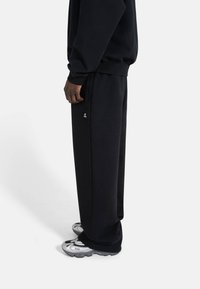 Svart joggebukse laget av myk stoff, med en avslappet passform, sidelommer, og en brodert logo nær midjen.