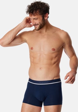 Blauwe katoenen boxershorts met een lichtgrijze tailleband, voorzien van een aansluitende pasvorm en een gladde textuur. Model toont een ontspannen houding.