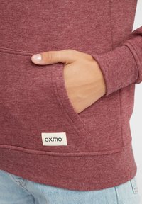 Bordo dukserica od mekanog materijala, s prednjim džepom i malim oznakom s natpisom "oxmo." Vidljive šavove duž šavova.