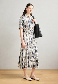 Shirt dress with a geometric pattern in navy and white, short sleeves, a belted waist, and paired with a black tote bag and beige flats.