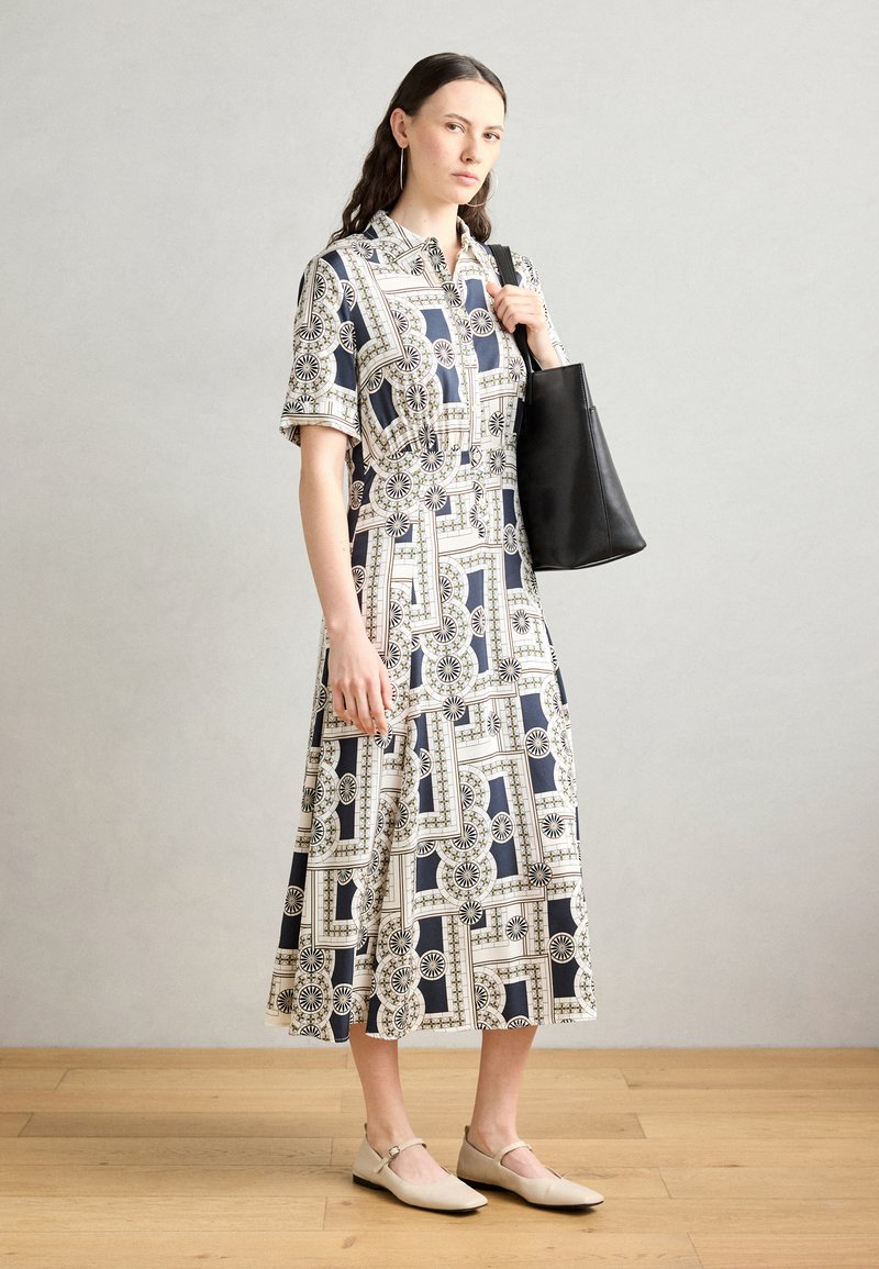 Shirt dress with a geometric pattern in navy and white, short sleeves, a belted waist, and paired with a black tote bag and beige flats.