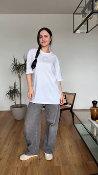 Giovane donna con capelli intrecciati indossa una maglietta bianca oversize, jeans grigi a gamba larga e sneakers, in piedi in un soggiorno moderno.