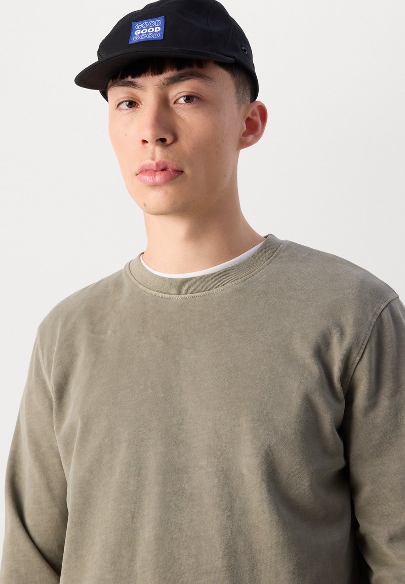 Sweatshirt vert olive avec un col rond, texture lisse et coupe décontractée ; associé à une casquette noire avec un patch logo bleu.