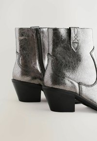 Botas metálicas plateadas hasta el tobillo con tacones gruesos negros, cremalleras laterales y un diseño decorativo en un fondo claro.
