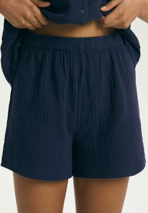 Osoba ubrana w granatowe szorty z teksturowanej tkaniny z elastycznym paskiem w talii, podciągająca dopasowaną granatową koszulkę, odsłaniającą brzuch.