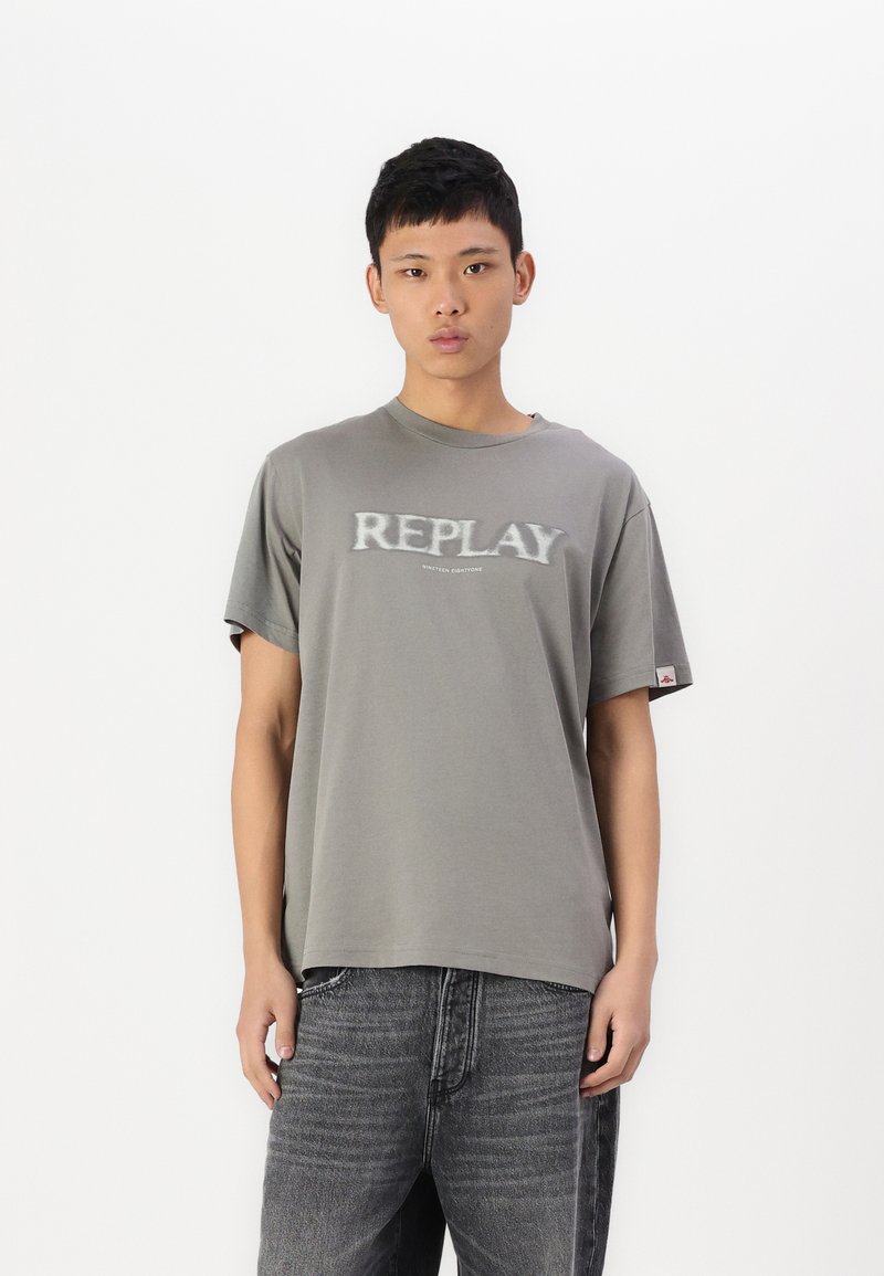 Grå bomulls t-skjorte med "REPLAY" i en litt uskarp trykk, kort ermer og en avslappet passform. Brukt med mørkegrå jeans.
