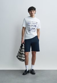 T-shirt en coton blanc avec motif de vague bleu, associé à un short foncé et des chaussures noires. Porte un sac à motif noir et blanc.