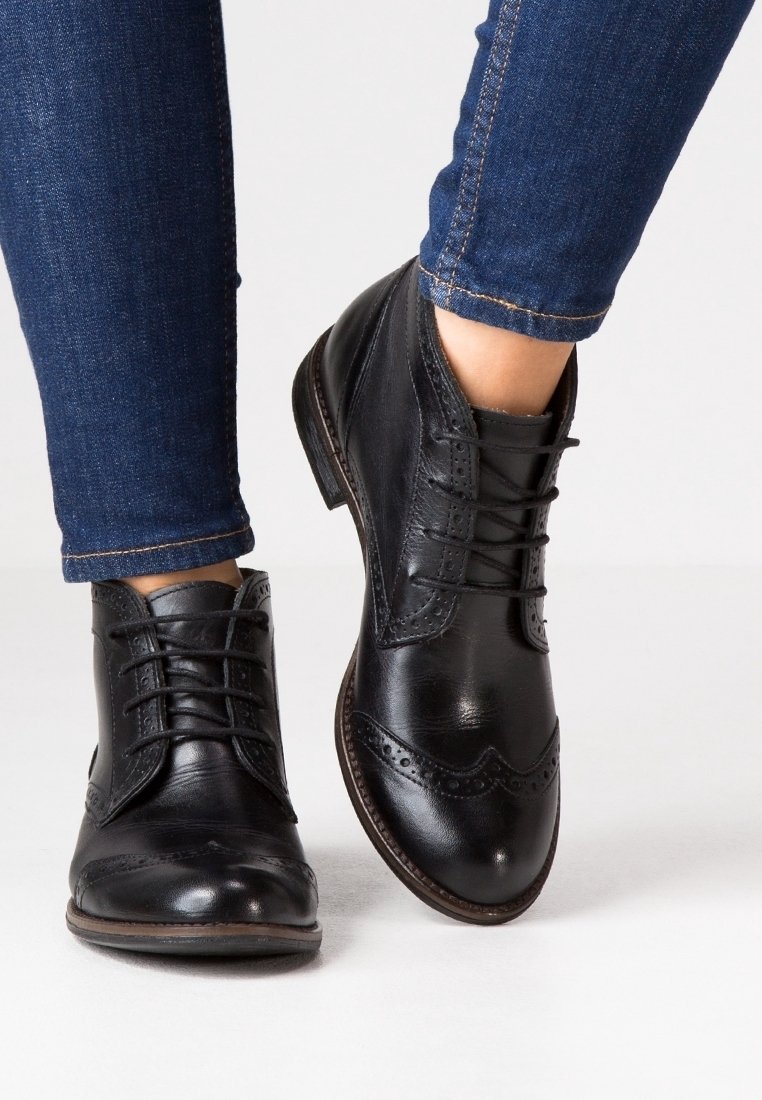 Botas de cuero negro hasta el tobillo con detalles de brogue, punta redonda y diseño con cordones. Combinadas con jeans azul oscuro, mostrando textura y contraste de color.