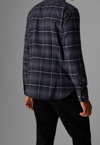 Chemise à carreaux sombre avec des lignes blanches et bleu clair, poignets boutonnés, ourlet arrondi, et texture du tissu visible à l'arrière.