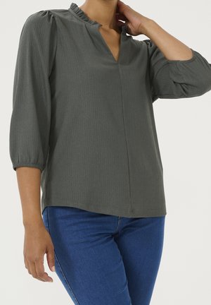 Blouse vert foncé avec un col à volants, des manches trois-quarts et un motif texturé subtil. Portée avec un jean bleu.