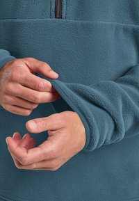 Fleece trui in teal met een half-rits ontwerp. Kenmerken zijn een zachte textuur, contrasterende geribbelde manchetten en aansluitende mouwen. Handen die de manchetten aanpassen.