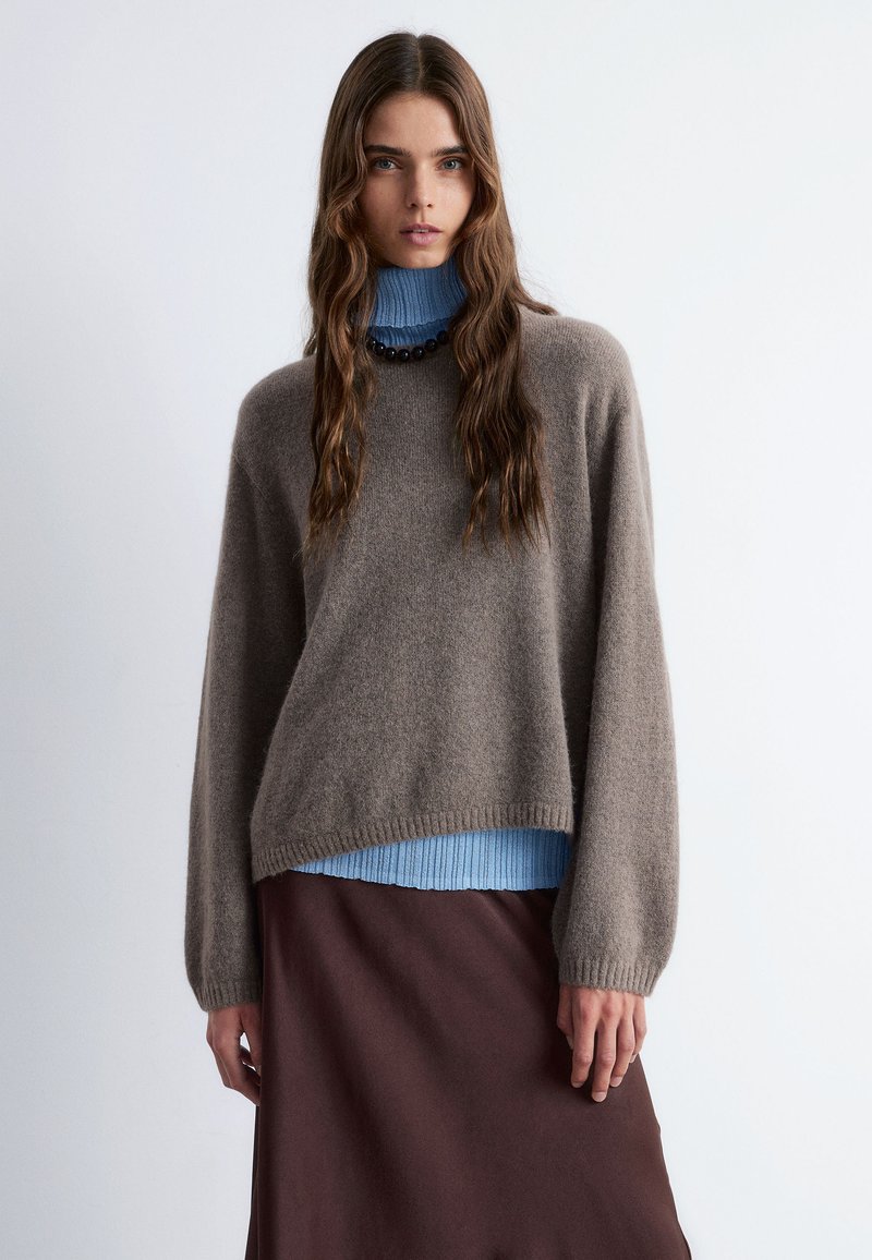 Brauner, gestrickter Pullover mit lockerer Passform, über einem hellblauen, gerippten Rollkragenpullover getragen, kombiniert mit einem dunkelbraunen Rock.