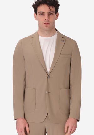 Uomo con capelli ricci scuri che indossa una giacca beige sopra una maglietta bianca a girocollo, in piedi contro uno sfondo semplice.