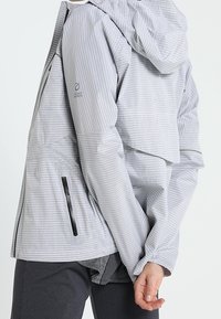 Jasnoszara, teksturowana kurtka z kapturem, z czarną kieszenią na zamek, noszona na ciemnych spodniach, osoba poprawiająca rękaw lewą ręką, trzymaną za plecami.