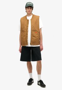 Chaleco acolchado marrón con cremallera frontal y dos bolsillos laterales, llevado sobre una camiseta blanca y combinado con pantalones cortos negros y zapatillas.