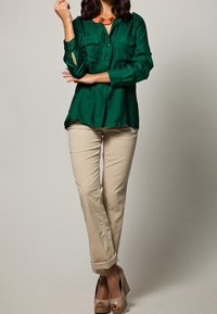 Grön satin skjorta med knapp-up och två bröstfickor. Tillsammans med ljusbeiga uppvikta byxor och tan plattformsskor. Accenthalsband synligt.