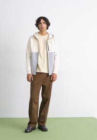 Muška lagana jakna na zip u off-white i sivoj boji, s kapuljačom i toniranim akcentima, u kombinaciji s smeškim hlačama i crnim obućom.
