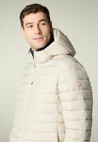 Uomo che indossa una giacca a vento trapuntata di un beige chiaro con cappuccio, mentre guarda di lato su uno sfondo neutro e semplice.