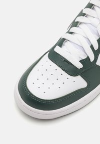 Vit och mörkgrön sneakerskaft med perforeringar, vita snören och texturerad sula på en enfärgad vit bakgrund.