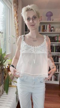 Lühikeste blondide juustega inimene kannab valget pitsist korsetti ja helesiniseid teksaseid, seistes siseruumis akna ja raamatukapi lähedal.